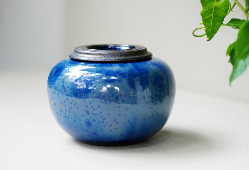 K&ouml;nnte beinhalten: Eine blaue Keramikdose mit einem dunkelgrauen Deckel. Die Dose hat eine gesprenkelte Glasur mit einem wirbelnden Muster.