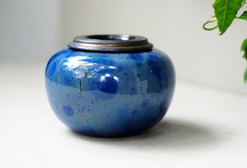 K&ouml;nnte beinhalten: Eine kleine, runde, blaue Keramikdose mit einem silbernen Deckel. Die Dose hat ein gesprenkeltes, marmoriertes Design.