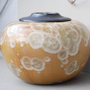 May include: A round, ceramic jar with a lid. The jar is a warm, golden-yellow color with white, circular patterns. The lid is a dark, muted blue. The jar has a glossy finish and appears to be handmade.