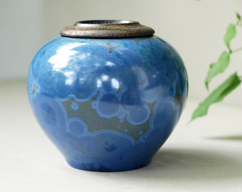 Op de afbeelding: Een blauwe keramische urn met een bruin deksel. De urn heeft een uniek patroon van blauwe en bruine cirkels.