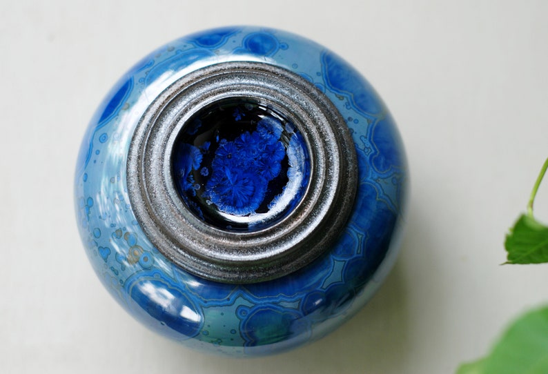 K&ouml;nnte beinhalten: Eine blaue Keramikdose mit einem silbernen Deckel. Die Dose hat ein wirbelndes Muster und ein blaues und schwarzes Inneres.