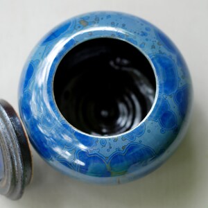 K&ouml;nnte beinhalten: Eine blaue Keramikdose mit Deckel. Die Dose hat ein wirbelndes Muster aus Blau und Wei&szlig;. Der Deckel ist schwarz und hat einen erh&ouml;hten Rand.