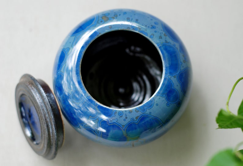 K&ouml;nnte beinhalten: Eine blaue Keramikdose mit Deckel. Die Dose hat ein wirbelndes Muster aus Blau und Wei&szlig;. Der Deckel ist schwarz und hat einen erh&ouml;hten Rand.
