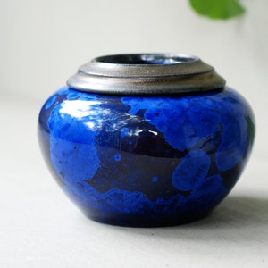 Può includere: Un piccolo vaso rotondo in ceramica blu con un coperchio in metallo argentato. Il vaso ha una smaltatura blu screziata con accenti blu scuro.