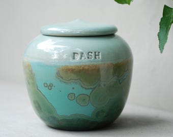 Op de afbeelding: Een keramische urn in turquoise, groen en bruin met een deksel. Het woord "DASH" is in reliëf op de voorkant aangebracht. De urn heeft een afgeronde vorm met een glanzende afwerking en de kleuren vloeien samen in een gemarmerd effect. De urn is waarschijnlijk voor as.