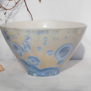 Op de afbeelding: Een witte keramische kom met een lichtblauwe en gele glazuur. De glazuur heeft een gespikkeld patroon met kleine blauwe cirkels.