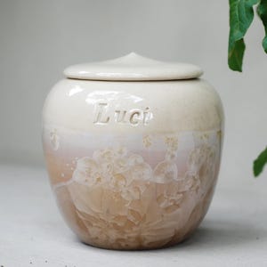 Op de afbeelding: Een keramische urn met een deksel, met een crème- en perzikglazuur met kristallijne patronen. De naam "Luci" is op de voorkant gegraveerd. De urn is ontworpen voor het bewaren van as en heeft een afgeronde vorm.