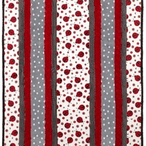 May include: A rectangular blanket featuring a repeating pattern of red ladybugs on a white background. The design includes vertical stripes of red, gray with white polka dots, and black patterned borders. The blanket has a black border.