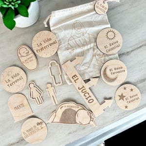 May include: Wooden craft pieces with Spanish text, including "La Vida Terrenal", "El Jucio", and "La Vida Preterrenal". The pieces are arranged on a light wood surface.