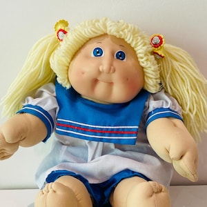 Puede incluir: Muñeca vintage con cabello rubio de lana en coletas, adornada con pequeños accesorios para el cabello amarillos y rojos. La muñeca tiene ojos azules, mejillas rosadas y viste un atuendo azul y blanco de estilo marinero.