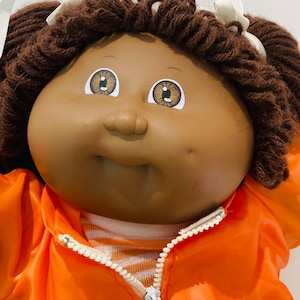 May include: A vintage Cabbage Patch Kids doll with brown yarn hair, brown eyes, and a tan complexion. The doll is wearing an orange jacket with a white zip and a striped shirt underneath. The Cabbage Patch Kids logo is visible on the jacket.