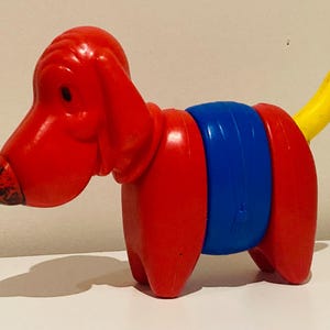 Può includere: Un giocattolo a forma di cane, rosso, blu e giallo. Il cane è fatto di plastica e ha la testa e le zampe rosse, una sezione centrale blu e una coda gialla. Il giocattolo è per bambini.