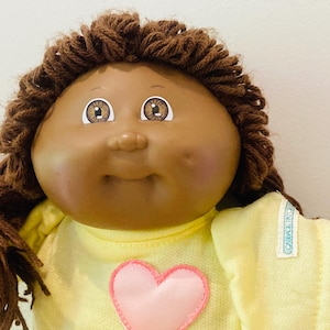 Peut inclure: Une poupée Cabbage Patch Kids vintage avec des cheveux bruns tressés. La poupée a les yeux bruns et une expression souriante. Elle porte une chemise jaune avec un cœur rose appliqué.