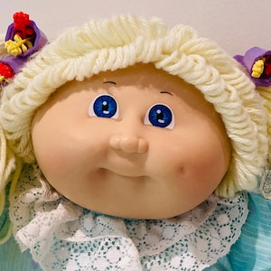 May include: A vintage Cabbage Patch Kids doll with a fair complexion, blue eyes, and a slight smile. The doll has light blonde yarn hair styled in pigtails, adorned with purple and red floral hair accessories. The doll is wearing a blue dress with a white lace collar.