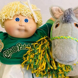 May include: A Cabbage Patch Kids doll in a green cheerleader outfit with a matching pom-pom, next to a plush grey donkey. The doll has blonde yarn hair and blue eyes. The donkey has a green and yellow rope halter.