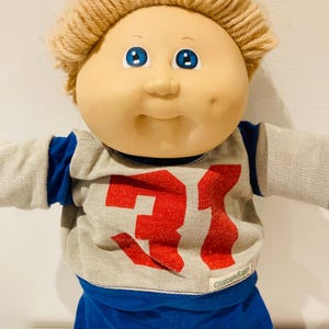 以下が含まれることがあります： 明るい肌色、青い目、ブロンドの糸の髪を持つ、ヴィンテージのキャベツパッチキッズ人形。人形は、赤で31の数字が入ったグレーとブルーのアメリカンフットボールのジャージと、青いショートパンツを着ています。クラシックなコレクターズアイテム。