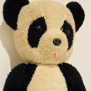 Può includere: Un peluche vintage a forma di panda. Il corpo è color crema con dettagli neri su orecchie, occhi e arti. Il panda ha un naso nero e un piccolo dettaglio rosso sulla bocca. Il peluche è realizzato in un materiale morbido e strutturato.
