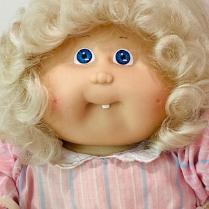 May include: A vintage doll with a round face, blue eyes, and a small, open mouth revealing two teeth. The doll has curly, light blonde hair and is wearing a pink and white striped dress with a white lace collar. A classic collectible toy.