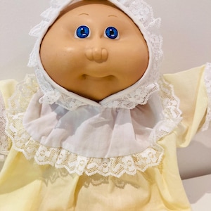 Pode incluir: Boneca Cabbage Patch Kid vintage com rosto redondo, olhos azuis e nariz e boca pequenos. A boneca está vestida com um vestido amarelo e um capuz branco com acabamento em renda.