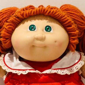 Puede incluir: Una muñeca vintage con coletas de hilo naranja y ojos verdes. La muñeca tiene tez clara, mejillas rosadas y un vestido rojo con cuello de encaje blanco. La cara de la muñeca tiene una expresión suave.