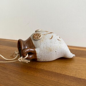 May include: A handmade ceramic hanging planter with a brown and white speckled finish. The planter has a small handle and a decorative spiral design. A natural fiber cord is attached for hanging.