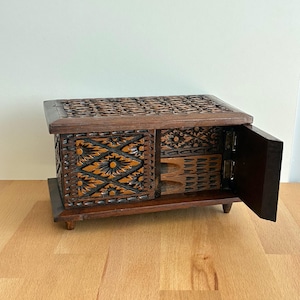 Può includere: Un portagioie in legno marrone scuro con intricati motivi floreali e geometrici intagliati. La scatola ha un coperchio incernierato e una piccola porta aperta, che rivela piccoli cassetti all'interno. La scatola è su una superficie di legno.