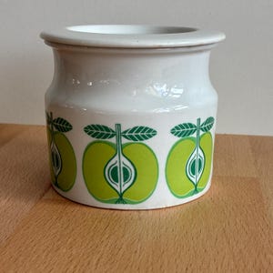 May include: White ceramic jar with a lid, decorated with a repeating pattern of green apple halves. The apples have green leaves and stems. The jar sits on a wooden surface.