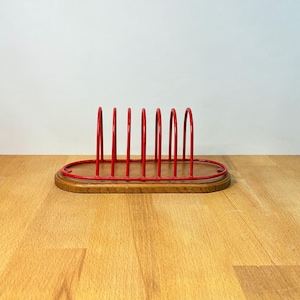 Puede incluir: Un soporte de alambre metálico rojo con seis divisores verticales arqueados unidos a una base de madera ovalada. La estructura presenta un acabado rojo liso y brillante en el marco de alambre y un acabado de veta de madera natural.