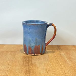 May include: A ceramic mug featuring a cylindrical body with a glossy blue glaze that transitions into a warm reddish-brown tone at the base. The mug includes a curved handle in a matching reddish-brown finish, showcasing a smooth, glazed texture.