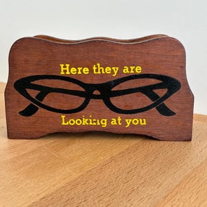 Puede incluir: Un soporte de gafas de madera con acabado marrón oscuro. El soporte presenta un gráfico de gafas negro y el texto amarillo "Here they are Looking at you". El soporte tiene una parte superior curva y está sobre una superficie de madera.