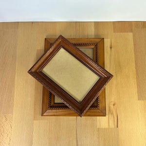 Pode incluir: Dois porta-retratos retangulares de madeira com acabamento em marrom médio e um padrão de borda decorativo estilo corda. As molduras estão empilhadas em ângulo, com uma posicionada diagonalmente sobre a outra, ambas com fundo bege liso.