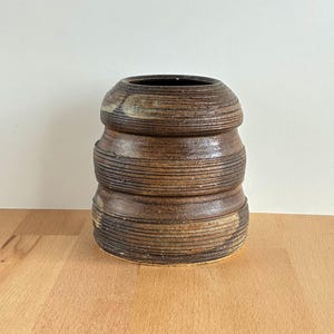 May include: A handmade ceramic vase with a stacked, cylindrical design. The vase has horizontal, textured lines and a dark brown colour with lighter brown and tan accents. The interior is dark. The vase sits on a wooden surface.