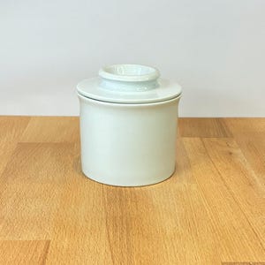 Könnte beinhalten: Ein massiver weißer Keramik-Buttertopf mit zylindrischer Basis und passendem Deckel, der einen mittigen Knaufgriff besitzt. Der Behälter hat eine glatte, glänzende Oberfläche und ein schlichtes, funktionales Design mit geraden Seiten.