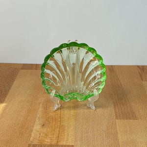 May include: A clear glass dish shaped like a scallop shell with a vibrant green rim. The dish features vertical ridges radiating from the base to the scalloped edge and is supported by a clear, three-legged plastic display stand for upright viewing.