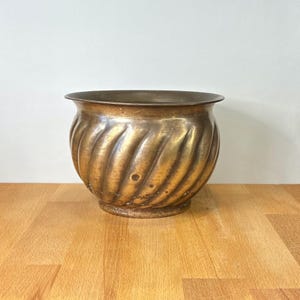 May include: A round brass planter featuring a distinct spiral fluted pattern along its exterior surface. The metal has a warm, aged golden patina with subtle variations in tone and a smooth, wide rim at the top, creating a classic bowl shape.