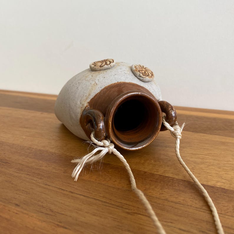 May include: A handmade ceramic hanging planter with a speckled white and brown finish. The planter has a wide opening and two small handles with attached rope for hanging. Two decorative floral accents are on the top.
