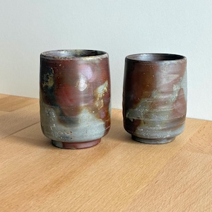 以下が含まれることがあります： 手作りの陶器のカップが2つ。カップは円筒形で、縁がわずかに広がっています。色合いは茶色、赤、グレーで、質感のある釉薬仕上げです。飲み物に適したカップです。