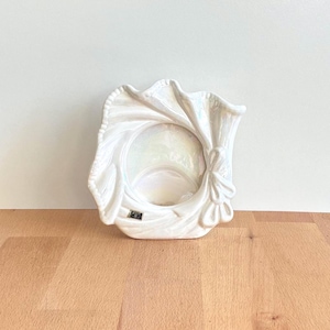 May include: A white ceramic photo frame with an iridescent finish, featuring a draped fabric design. The frame has a circular aperture and a decorative bow. It sits on a wooden surface.