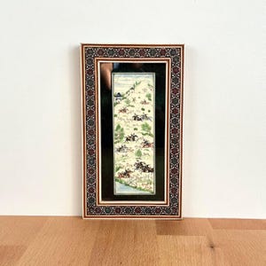 Puede incluir: Una pintura en miniatura enmarcada que representa una escena de caza con figuras a caballo, perros y fauna. La obra de arte está ambientada en un paisaje con árboles y agua. El marco tiene un borde decorativo con detalles en rojo, marrón y crema.