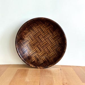 May include: A circular wooden tray in dark brown, featuring a woven design. The tray's edge is slightly raised, and the pattern is created by alternating light and dark brown wood strips. The tray's diameter is approximately 30 cm.