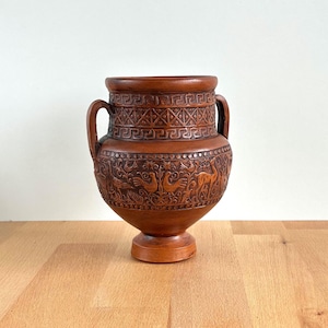Puede incluir: Un jarrón de cerámica marrón con dos asas y una base. El jarrón presenta intrincados relieves de patrones geométricos, pájaros y ciervos. El jarrón se exhibe sobre una superficie de madera.