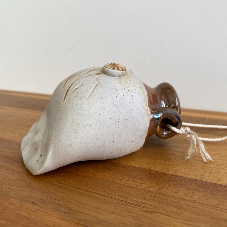 May include: A handmade ceramic bell with a cream-colored body and a brown spout. The bell features carved lines and a small, decorative element on top. A natural fiber cord is attached for hanging. The bell is likely for home decor.