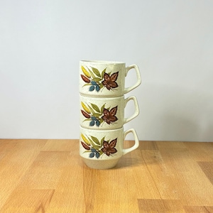May include: A stack of three cream-colored ceramic mugs with a speckled finish. Each mug features a botanical illustration of brown maple leaves, green foliage, yellow wheat stalks, and small blue berries on the front side with a handle.