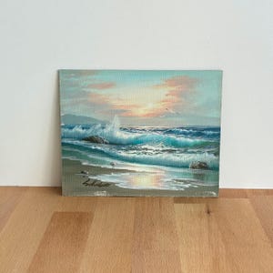 Puede incluir: Pintura al óleo de un paisaje marino con olas turquesas rompiendo, una playa de arena y una puesta de sol con tonos rosas y naranjas. Dos gaviotas vuelan en el cielo. La pintura está firmada en la esquina inferior izquierda.