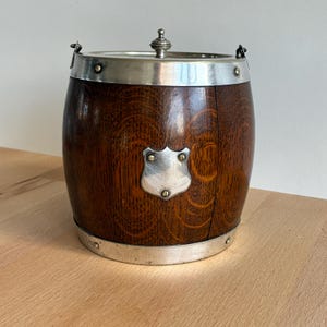 May include: A wooden barrel-shaped ice bucket with a silver-colored rim and handle attachments. The dark brown wood has a visible grain pattern. A silver shield-shaped emblem is affixed to the front. The lid is also silver-colored and has a decorative knob.