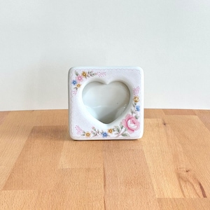 May include: White ceramic heart-shaped photo frame with floral detailing. The frame features a heart-shaped aperture and is adorned with pink, blue, and yellow flowers. The square frame has a scalloped border.