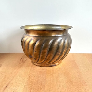 May include: A vintage brass planter with a spiral design. The planter has a wide, rounded shape with a slightly flared rim. The brass has a warm, aged patina, adding to its antique charm. The planter sits on a wooden surface.