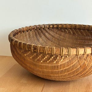 Pu&ograve; includere: Un cesto rotondo intrecciato in tonalit&agrave; marroni naturali. Il cesto presenta un design ampio e aperto con un bordo rinforzato. L'intreccio intricato crea una superficie strutturata. Probabilmente realizzato in rattan o materiale simile.