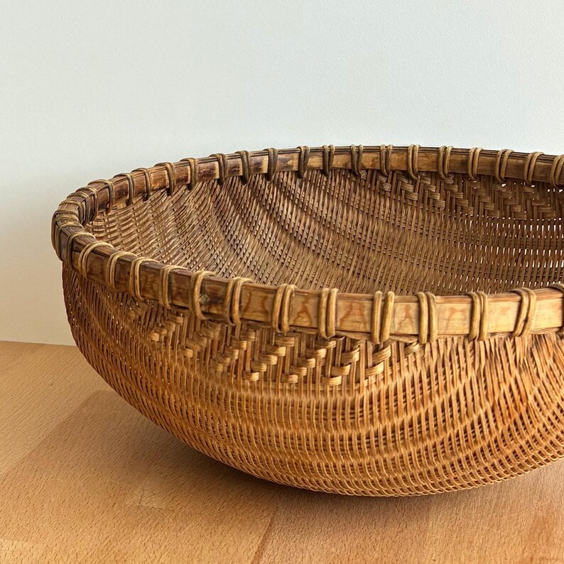 Pu&ograve; includere: Un cesto rotondo intrecciato in tonalit&agrave; marroni naturali. Il cesto presenta un design ampio e aperto con un bordo rinforzato. L'intreccio intricato crea una superficie strutturata. Probabilmente realizzato in rattan o materiale simile.