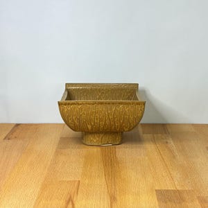 May include: A square ceramic bowl with a mustard yellow glaze and a textured, raised pattern resembling tree bark or organic veins. The bowl features a wide, open top with straight edges and sits on a raised, square pedestal base.
